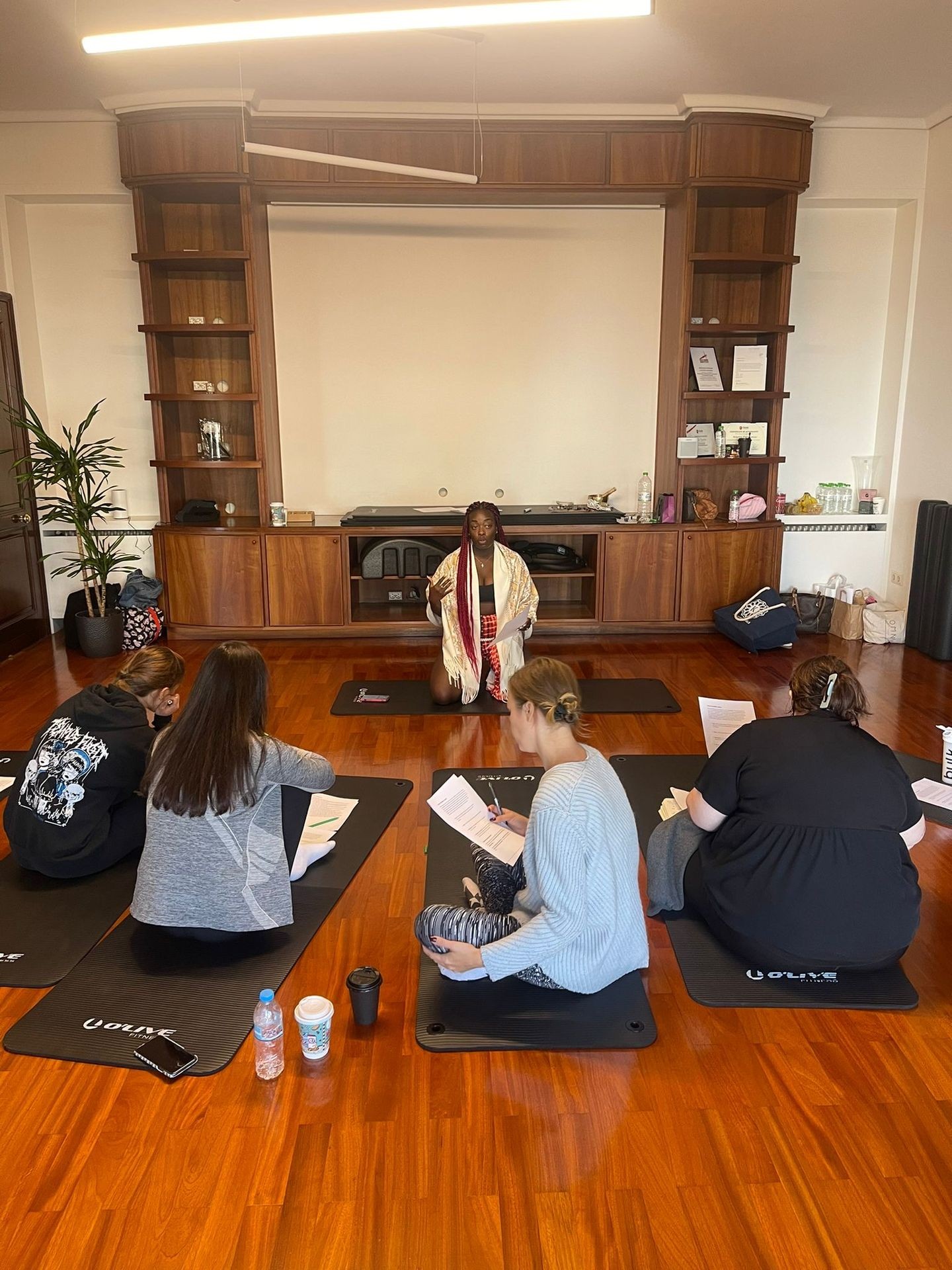 Group meditation session with instructor on a wooden floor, participants seated on mats holding papers.